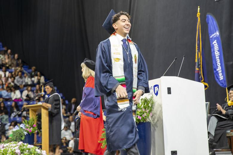 Graduate Rogelio Flores wears cap, gown, and Mexican heritage sash while crossing stage 